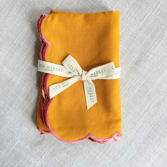 A folded mustard yellow fabric napkin with pink scalloped edges, tied with a cream ribbon.