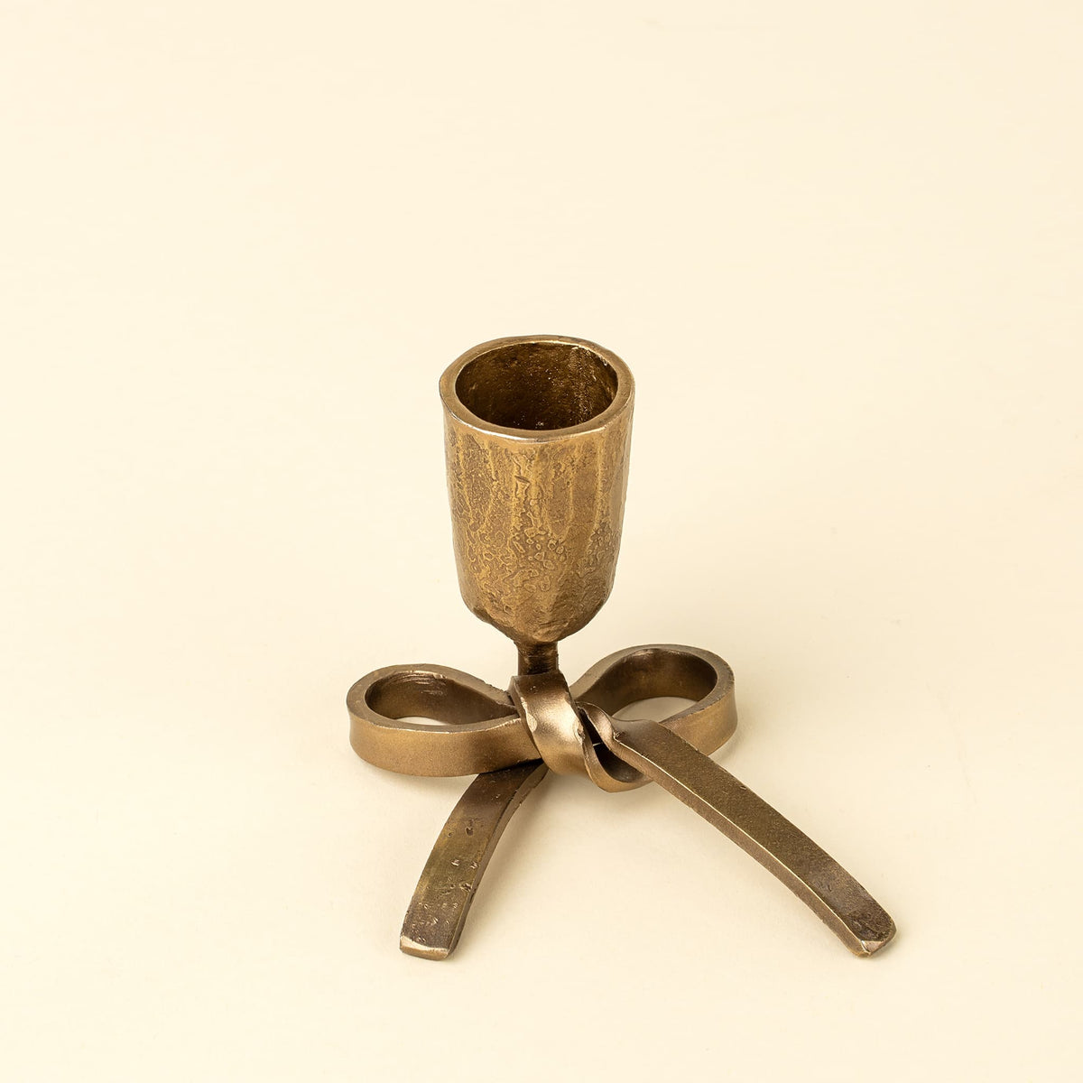 A small metallic candle holder with a textured cylindrical cup and a bow-shaped base, shown on a plain warm cream background.