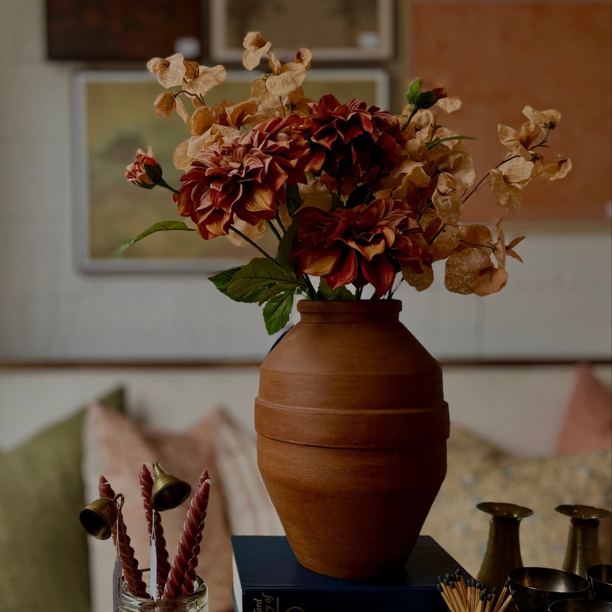 A terracotta-coloured ceramic vase filled with dried orange, brown, and beige flowers, styled on a dark blue book in a cozy, dimly lit room with soft-focus background decor.