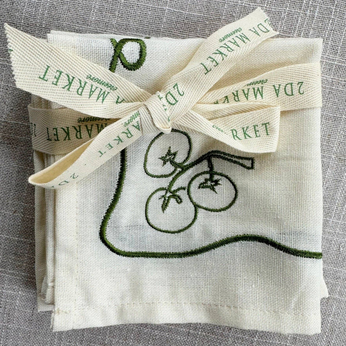 A folded cream-colored linen cloth with green embroidered tomato motifs and a matching ribbon tied around it, placed on a textured gray fabric background. 