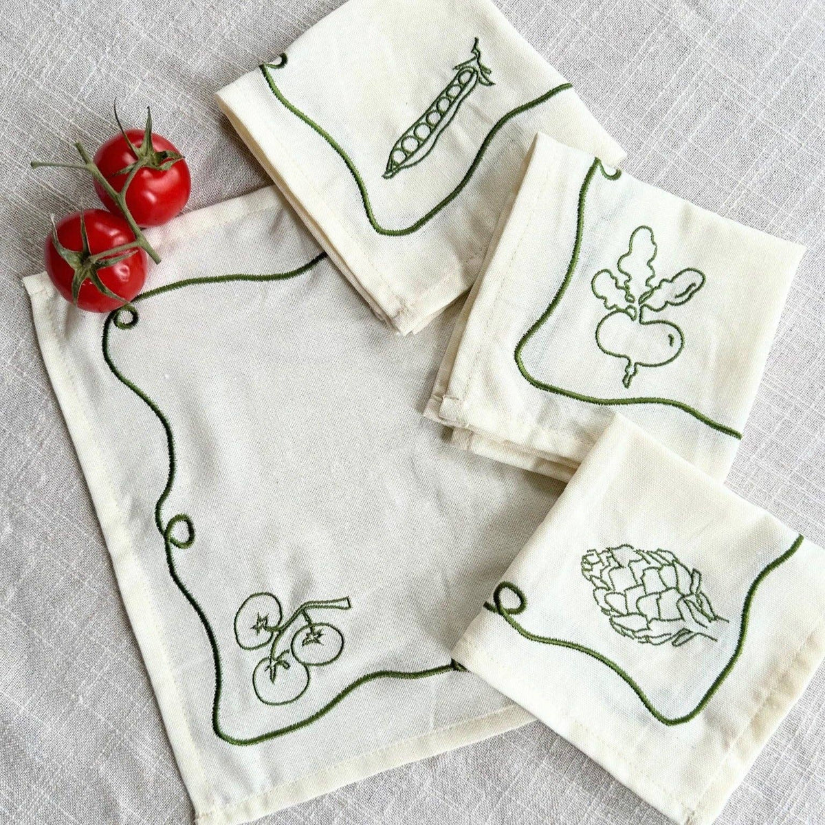 Four cream-colored linen cocktail napkins with green embroidered vegetable motifs (pea pod, radish, tomatoes, hop) arranged on a textured light gray fabric background with two small red tomatoes on the side.