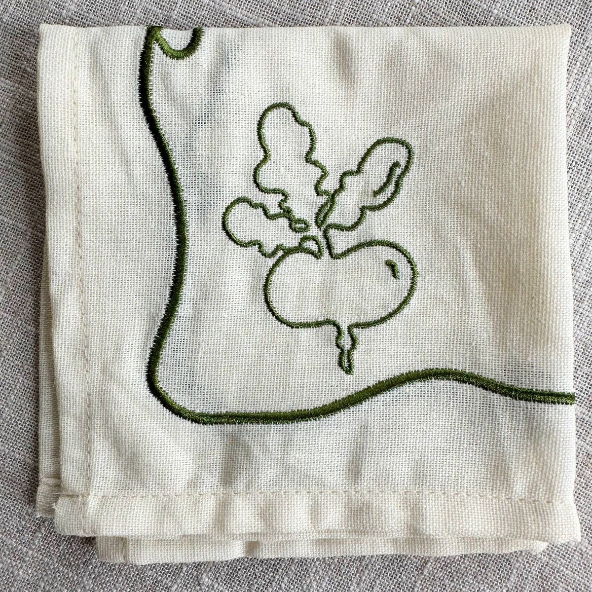 A folded cream-colored cotton kitchen towel with green embroidered details, featuring a simple outline of a radish on one corner and a curved green border.