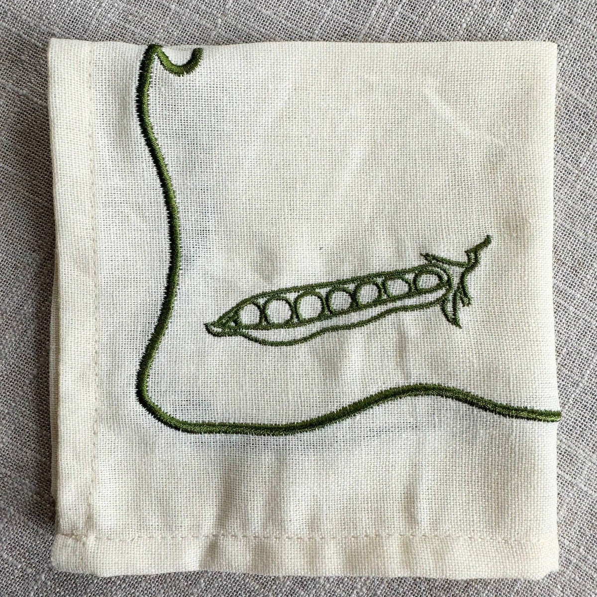 An off-white linen napkin featuring a green embroidered design of a pea pod and a curved vine, laid flat on a textured gray fabric background.