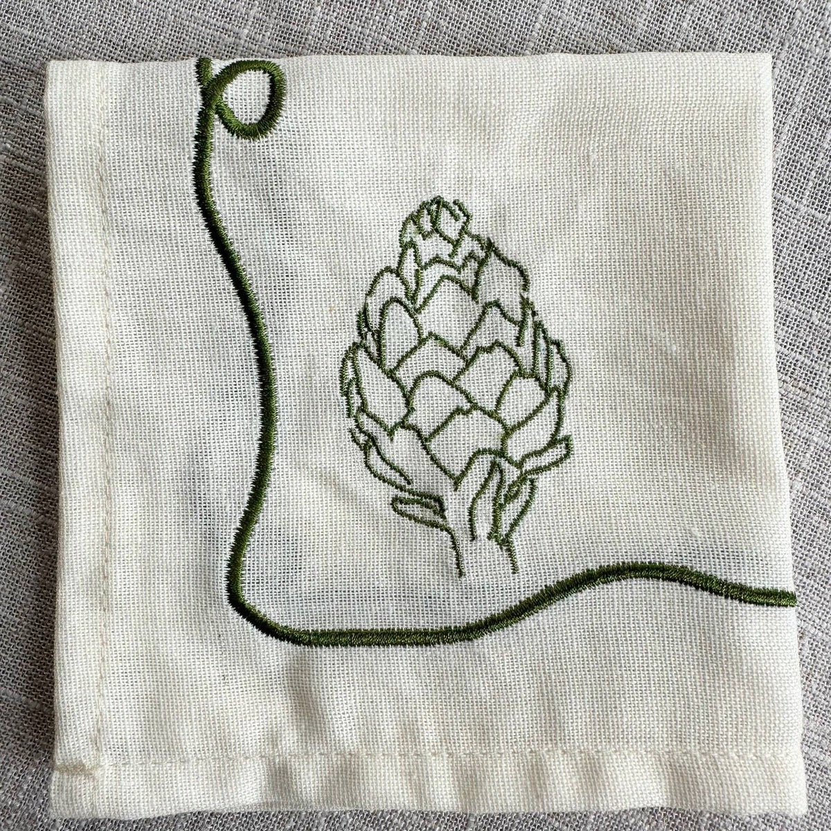 A square cream-colored cotton napkin with dark green embroidered details, featuring a stylized artichoke in the center and a decorative border, laid flat on a textured gray linen background.