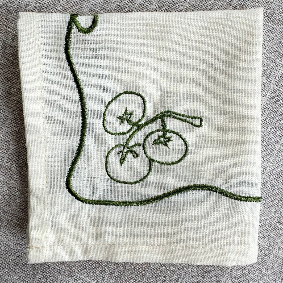 An off-white linen napkin with dark green embroidered tomatoes on a vine, laid flat on a textured gray linen surface.