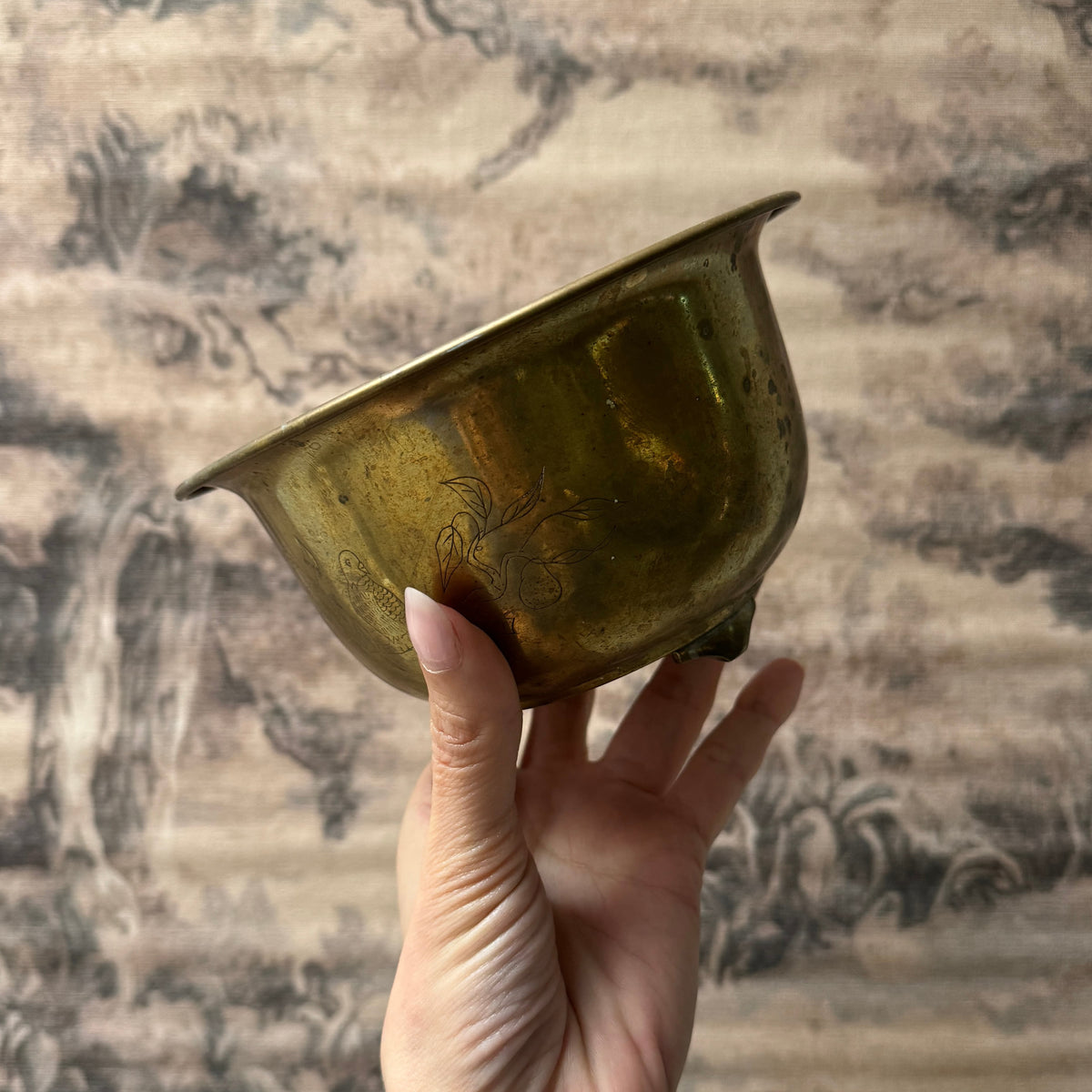 A hand holding a small, antique brass bowl with engraved details, set against a traditional landscape tapestry backdrop.